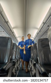 Young Pretty Airline Hostesses In Blue Uniforms Standing In The Aircraft Cabin And Smiling Friendly