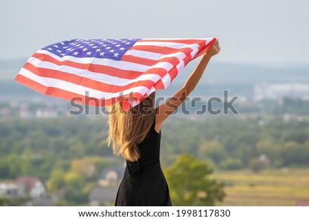 Young pretty american woman with long hair holding waving on wind USA  flag on her sholders standing outdoors enjoying warm summer day.