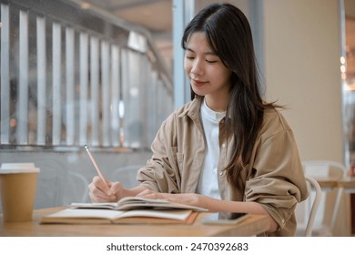 A young pretty Asian female college student in comfy clothes doing her homework at a cafe in the city, writing something in a notebook at a table. - Powered by Shutterstock