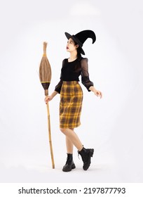 Young Pretty Asian Woman In Halloween Costume Wearing Witch Hat Holding Broom To Celebrate Halloween Party Posing On White Background.