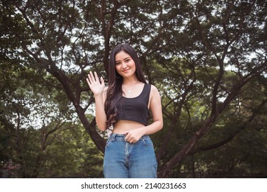 A Young Pretty Asian Woman Saying Hi. Waving To Someone In The Park. Approaching A Friendly Gesture.