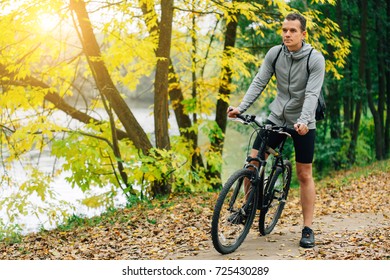 Young Pretty Athletic Man Standing With Bicycle In Colorful Autumn Park. Fall Season Background. Male Cyclist On The Road With Fallen Leaves