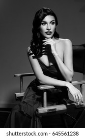 Young Pretty Beautiful Woman In Red Long Evening Dress With Makeup, Red Lips And Classical Hollywood Waves Hairstyle Sitting In Director's Chair At Film Set With Two Cinema Lights.