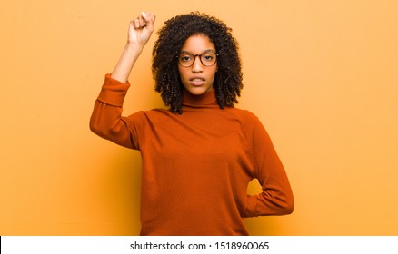 Young Pretty Black Woman Feeling Serious, Strong And Rebellious, Raising Fist Up, Protesting Or Fighting For Revolution Against Orange Wall