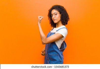 young pretty black woman feeling happy, satisfied and powerful, flexing fit and muscular biceps, looking strong after the gym against orange wall