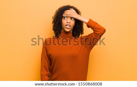 young pretty black woman panicking over a forgotten deadline, feeling stressed, having to cover up a mess or mistake against orange wall