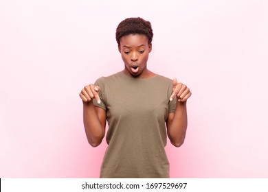 young pretty black womanfeeling shocked, open-mouthed and amazed, looking and pointing downwards in disbelief and surprise against pink wall