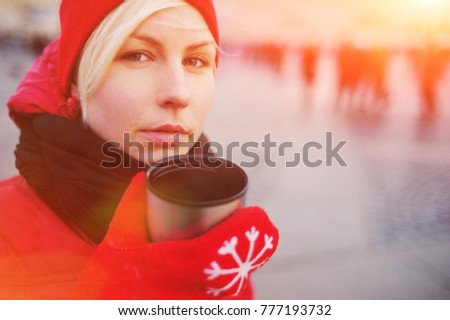 Young pretty blonde girl in a red dress holding a hand mittens a Cup of hot tea from a thermos in the winter while walking