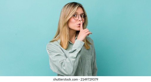 young pretty blonde woman asking for silence and quiet, gesturing with finger in front of mouth, saying shh or keeping a secret