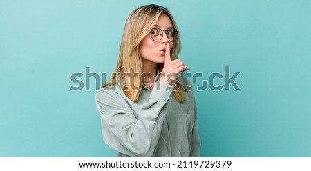 young pretty blonde woman asking for silence and quiet, gesturing with finger in front of mouth, saying shh or keeping a secret
