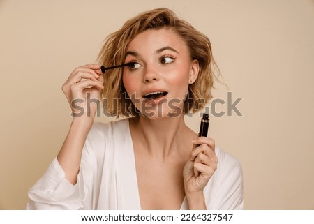 Young pretty blonde woman doing make up with mascara isolated over beige studio wall