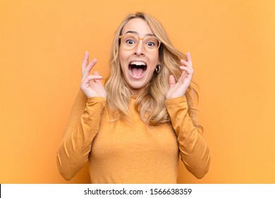 young pretty blonde woman feeling shocked   excited  laughing  amazed   happy because an unexpected surprise against flat color wall