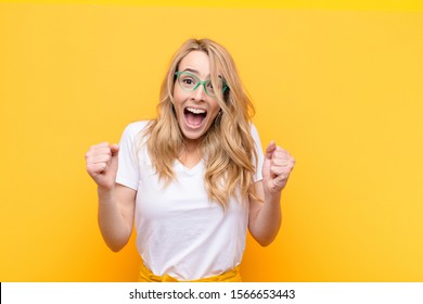 young pretty blonde woman feeling shocked  excited   happy  laughing   celebrating success  saying wow! against flat color wall