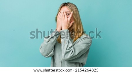 young pretty blonde woman feeling scared or embarrassed, peeking or spying with eyes half-covered with hands