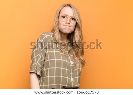 young pretty blonde woman with a goofy, crazy, surprised expression, puffing cheeks, feeling stuffed, fat and full of food against flat color wall