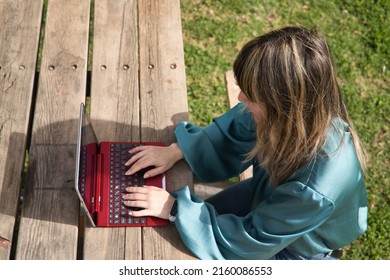 Young, Pretty, Blonde Woman In Green Shirt, Sitting On A Wooden Bench In A Park, Working With Her Laptop, View From Above. Technology Concept, Tablet, Notebook, Teleworking, Telecommuting, Zenithal.
