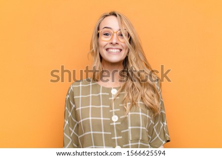 young pretty blonde woman looking happy and goofy with a broad, fun, loony smile and eyes wide open against flat color wall