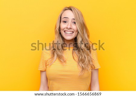 young pretty blonde woman looking happy and goofy with a broad, fun, loony smile and eyes wide open against flat color wall