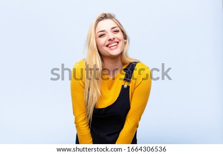 young pretty blonde woman looking happy and goofy with a broad, fun, loony smile and eyes wide open against flat wall