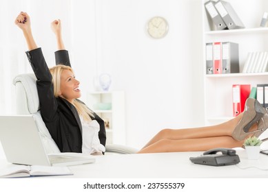 Young Pretty Blonde Woman In The Office Sitting In Office Chair, Hands Up And With Her Feet Up On A Desk. In The Background You Can See The Shelves With Binders.