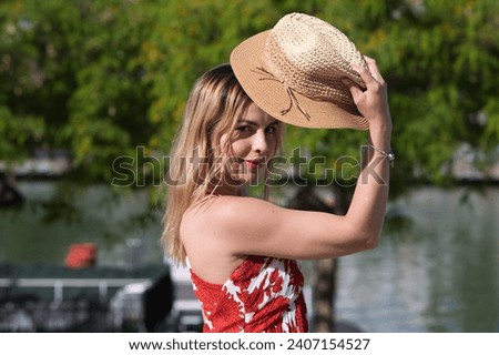 Young, pretty blonde woman takes straw hat off her head. She is dressed in a white dress with red print and is having fun doing different poses. In the background the river and trees