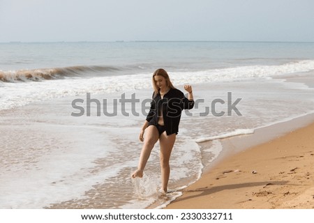 Young, pretty blonde woman walks along the shore of the beach wearing a black shirt and black bikini and plays at kicking in the water. In the background the sea and the sky.