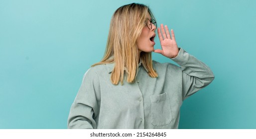 Young Pretty Blonde Woman Yelling Loudly And Angrily To Copy Space On The Side, With Hand Next To Mouth