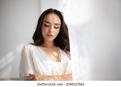 Young pretty brunette girl with incredible makeup in white summer dress among white tenderness tulle standing near the window. Indoor photography. - Shutterstock ID 2048167865