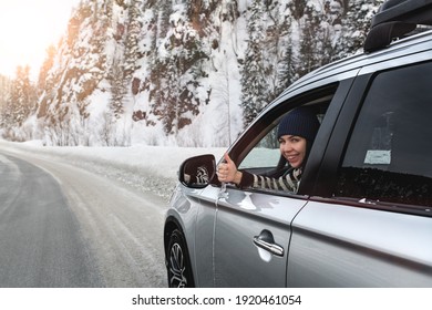 Young pretty brunette sitting in a car and showing thumb up sign through opened side window in sunny winter day on icy road close up.