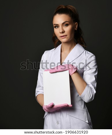 Young pretty brunette woman doctor therapist nurse in white uniform and protective gloves holding blank paper sheet over dark background, copy space. Medicine workers and healthcare concept