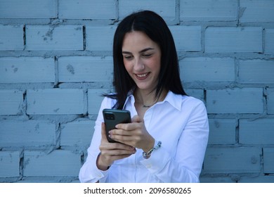 Young, pretty brunette woman is looking at her mobile phone. In the background a grey brick wall. Concept of new technologies and social networks.