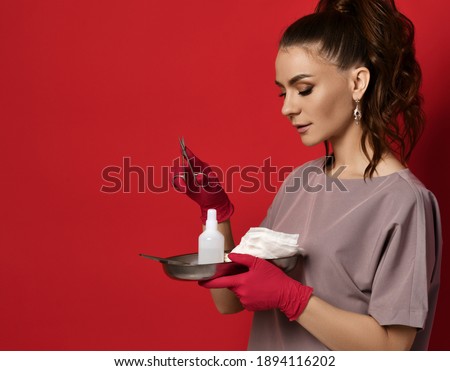 Young pretty brunette woman nurse doctor in latex gloves holds medical tools antiseptic bandage on tray and medical scissors in hand over red wall background. Gynecology, healthcare, medicare concept