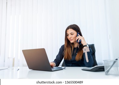 Young, Pretty Business Woman With Long Brown Hair Talking On The Phone While Sitting Behind Her Office Desk.