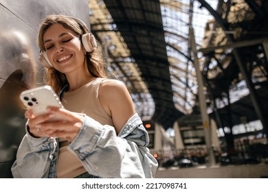 Young pretty caucasian girl waiting for train and listening to music from phone in big headphones. Blonde smiles, wears denim jacket. Gadgets concept - Powered by Shutterstock