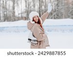 young pretty caucasian redhead smiling girl in white and beige winter clothes skating in ice rink in winter cloudy snowy day, with skates on shoulder, looking straight to camera, waving hand