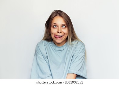 A Young Pretty Caucasian Ridiculous Blonde Woman In An Oversize T-shirt Shows Tongue Looking Up, Grimacing And Showing A Funny Face Isolated On A White Background