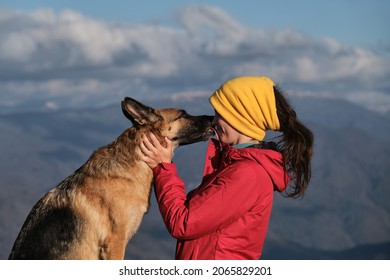 Young pretty Caucasian woman in red jacket stands on top of mountain next to her dog and enjoys scenery at sunset. Travel with German shepherd and owner. Hug and kiss best friend in nature.