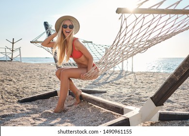 Young pretty cheerful blond woman in red swimsuit wearing sunglasses and hat sitting in hammock happily looking in camera on beach
