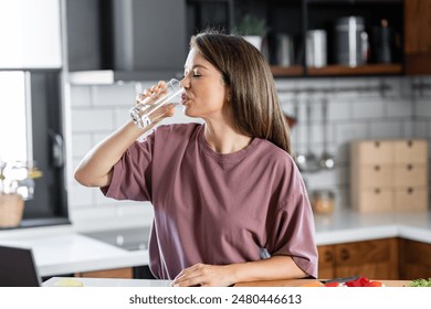 A young pretty cheerful woman is drinking a glass of water in home kitchen in the morning - Powered by Shutterstock