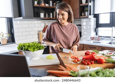 A young, pretty, cheerful woman is looking at a laptop computer while preparing food at home, following a cooking recipe online - Powered by Shutterstock