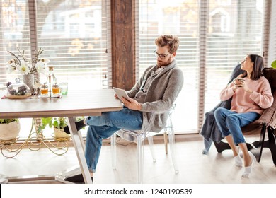 Young pretty couple in home clothes and slippers sitting at kitchen with large windows talking together about new project while drinking morning coffee and smartphone waiting for their friend to come. - Powered by Shutterstock