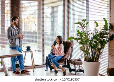 Young pretty couple in home clothes and slippers sitting at kitchen with large windows talking together about new project while drinking morning coffee and smartphone waiting for their friend to come. - Powered by Shutterstock