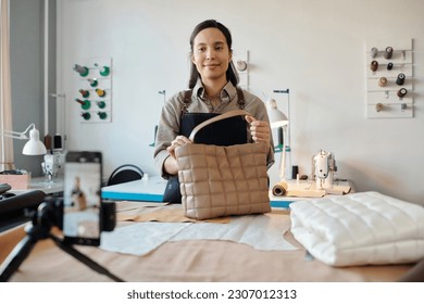 Young pretty craftswoman in workwear giving masterclass on leatherworking while looking at smartphone camera in studio - Powered by Shutterstock