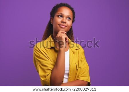Young pretty ethnic African American woman thinking holding hand on chin imagining happy future or coming up with new creative ideas dressed in stylish casual clothes stands on purple solid background