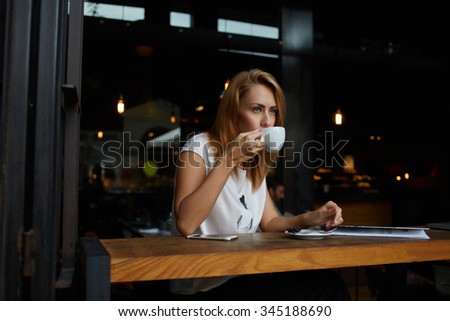Young pretty European woman drinking cappuccino while relaxing in modern cafe bar after work day, dreamy female enjoying cup of tea while sitting in cozy coffee shop during her recreation time 