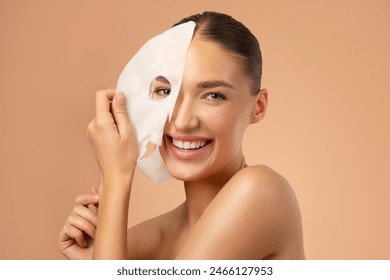 Young pretty European woman removing moisturizing, rejuvenating white fabric sheet mask from her face after using it, smiling at camera, beige background - Powered by Shutterstock