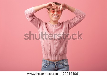 young pretty exited sincere laughing woman in pink sweater and sunglasses isolated on pink studio background, candid smile