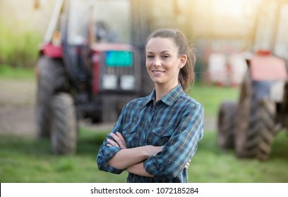 Young pretty farmer woman standing on farmland with crossed arms and tractors in background - Shutterstock ID 1072185284