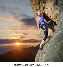 Young pretty female climber climbing with rope and carbines on large boulder against scenic sunset background. Hanging on one hand, smiling and looking at the camera. Summer time. 1x1 