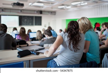 Young Pretty Female College Student Sitting In A Classroom Full Of Students During Class